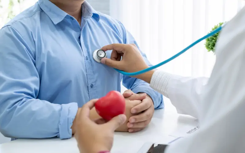 Un médecin ausculte un patient au stéthoscope ; un cœur en mousse rouge est posé sur le bureau.