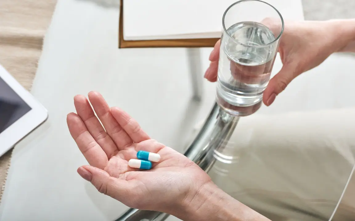 One hand holds out a glass of water above an open palm with two capsules in it.