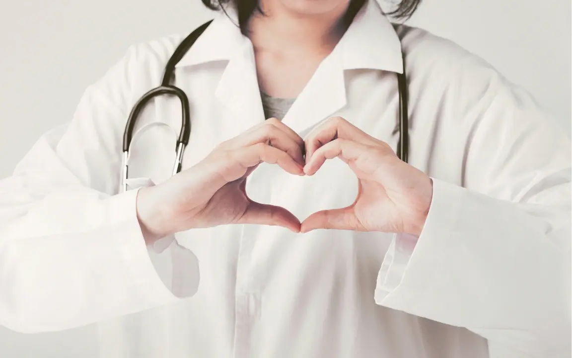 Une soignante en blouse blanche forme un cœur avec ses mains devant son stéthoscope.