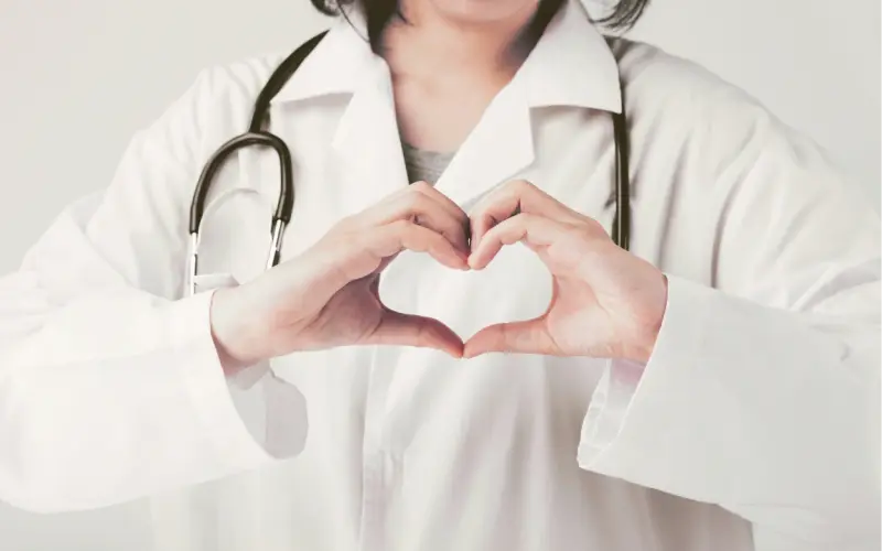 Une soignante en blouse blanche forme un cœur avec ses mains devant son stéthoscope.