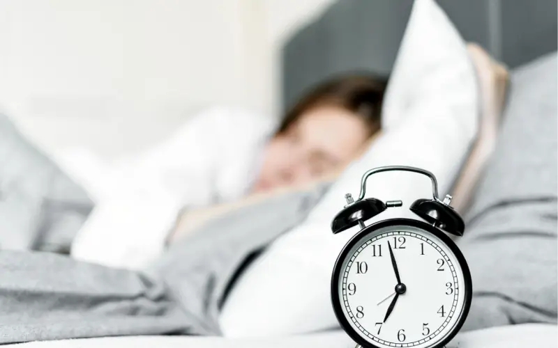 Une personne dort dans son lit, un réveil posé au premier plan.