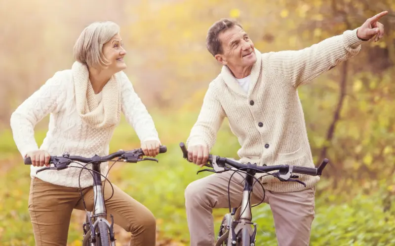 An older couple cycling through a park in autumn, one of them pointing into the distance.