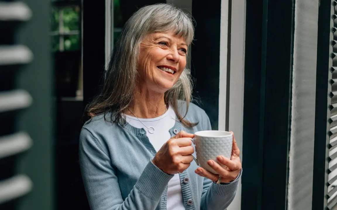 Une femme aux cheveux gris, une tasse entre les mains, regarde par la fenêtre de son salon.