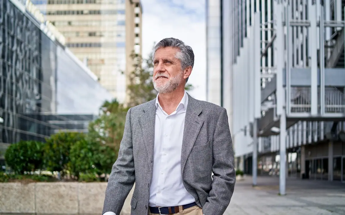 Un homme aux cheveux gris, veste sur les épaules, marche dans une rue devant un bâtiment moderne.