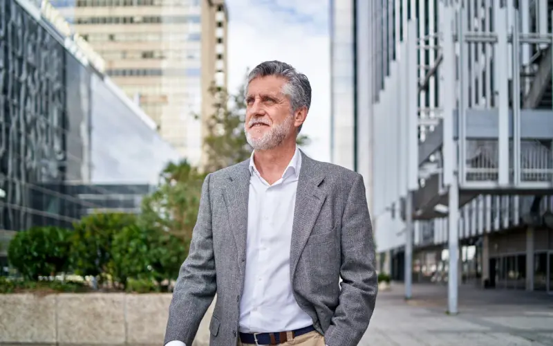 Un homme aux cheveux gris, veste sur les épaules, marche dans une rue devant un bâtiment moderne.