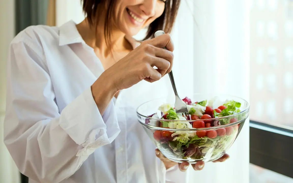 Une femme tient un saladier et pique une tomate cerise à la fourchette, devant une fenêtre.